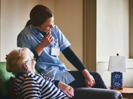 Young female nurse and elderly woman sitting on couch and smiling at care home